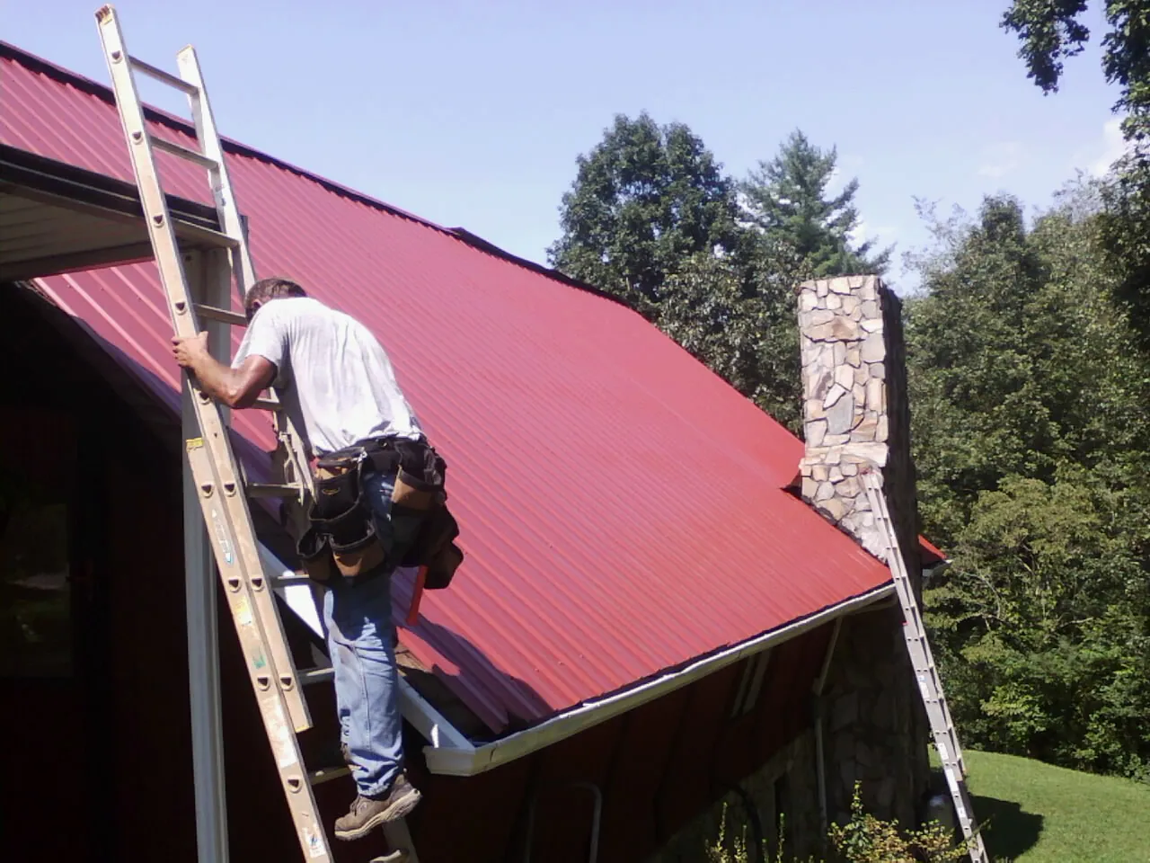 Roofer on a ladder beside a red metal roof and stone chimney — Soffit & Fascia Repair by Roofing Brothers in Jersey City